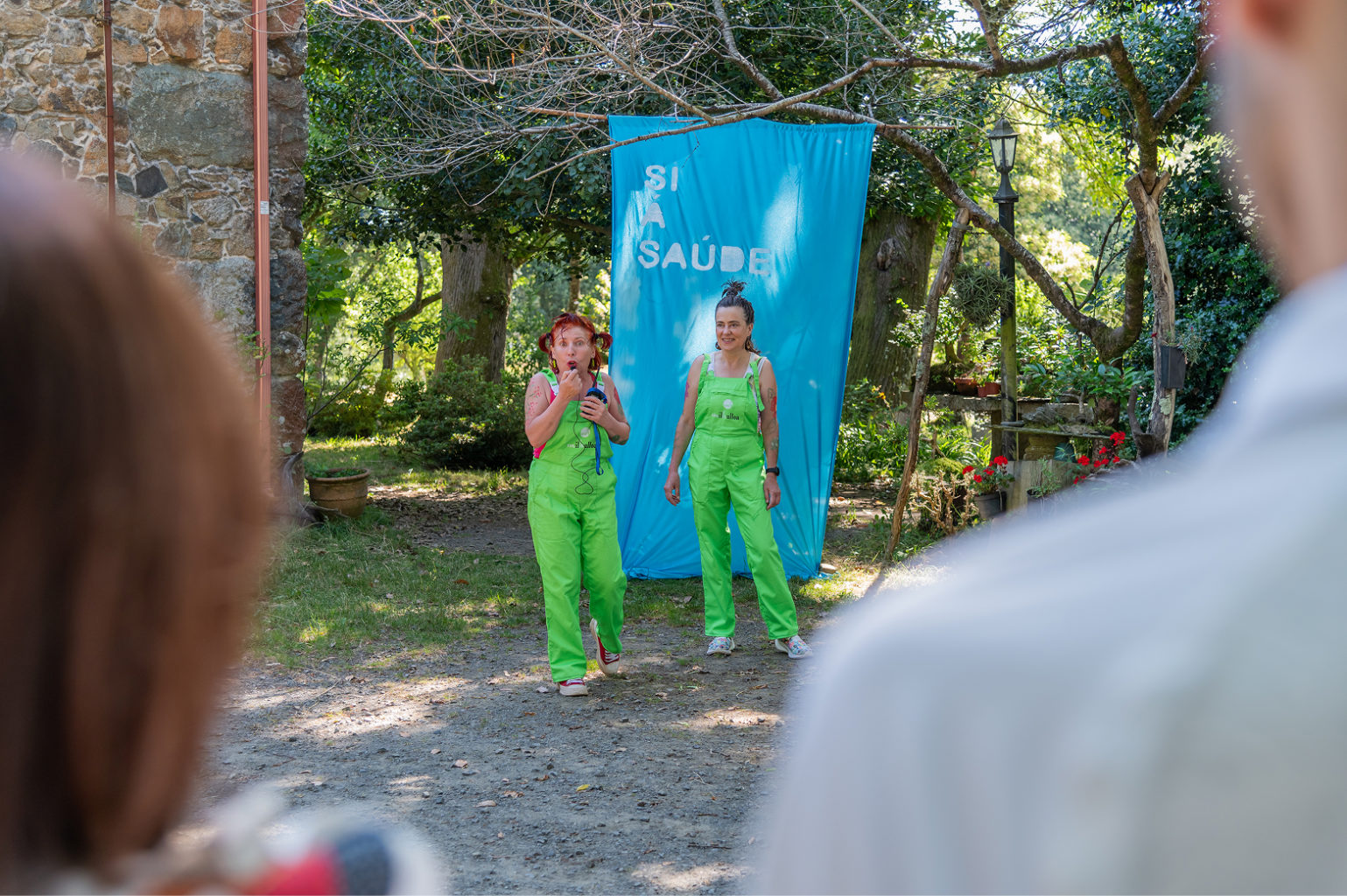 Two people talk to the public. Behind them a banner reads "Si á saúde".